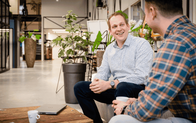 Two colleagues in an open office, talking by a low wooden table with a laptop and coffee cups.